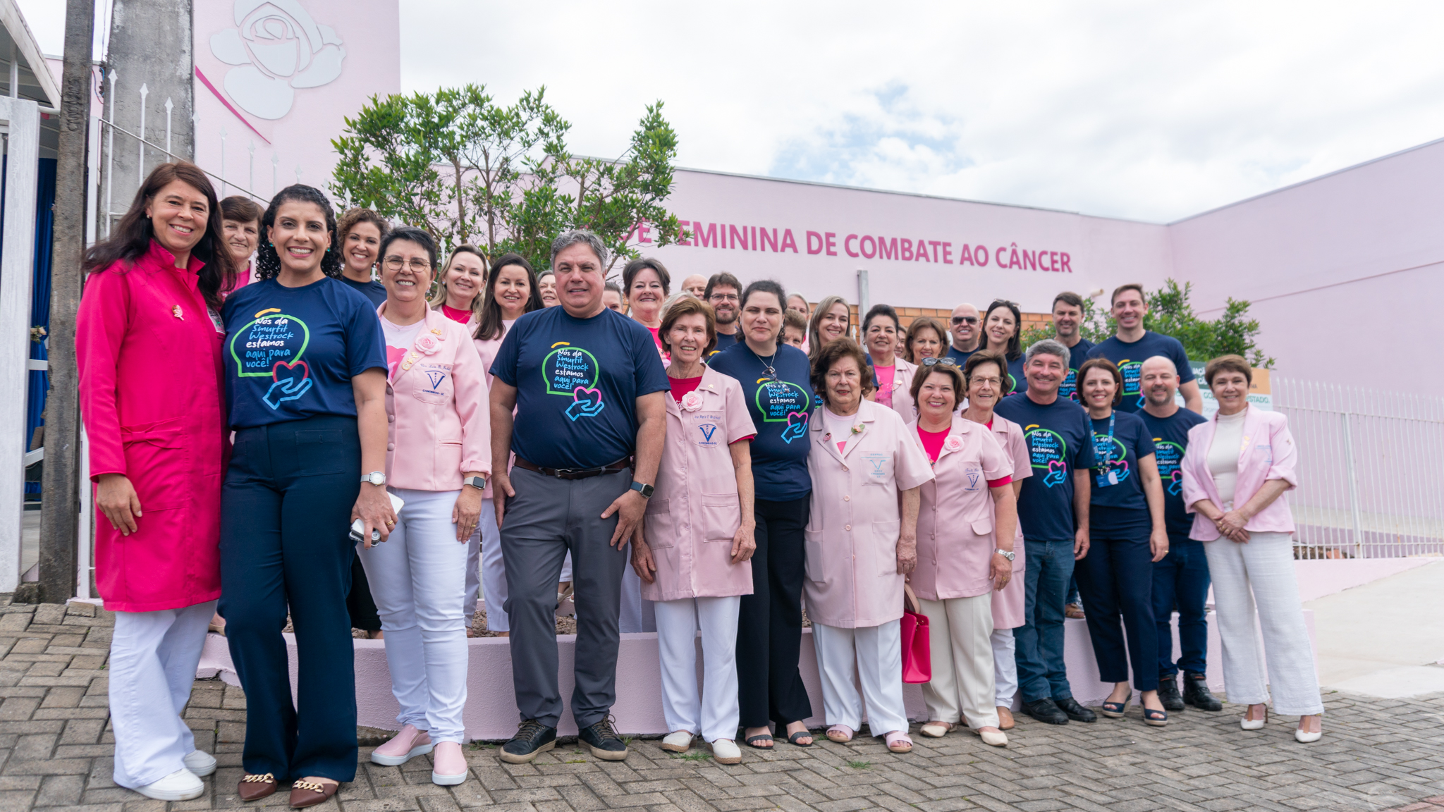 Grupo de colaboradores e voluntários reunidos em frente à Rede Feminina de Combate ao Câncer, durante ação social de apoio e conscientização.