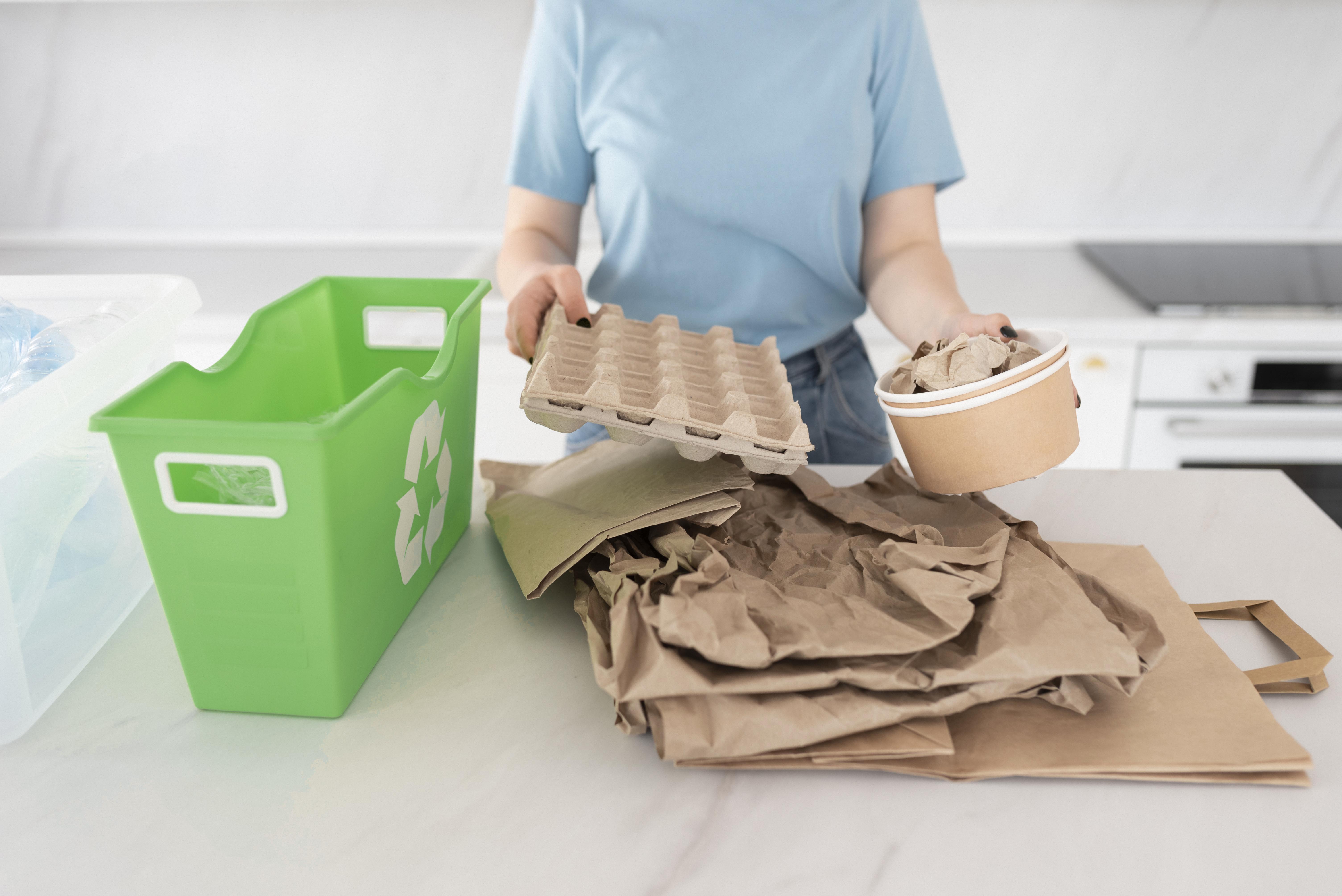 Pessoa separando embalagens de papel e materiais recicláveis ao lado de uma lixeira de reciclagem verde, representando práticas sustentáveis e descarte consciente de embalagens.