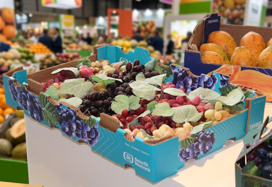 Fotografia em close de caixas de papelão personalizadas da Smurfit Westrock em uma feira comercial de frutas. Em primeiro plano, uma caixa azul vibrante está repleta de uvas verdes, rosadas e pretas, decoradas com folhas artificiais. Ao fundo, à direita, uma caixa azul escura está preenchida com mamões (papaias) maduros. O cenário ao redor é o de um pavilhão de eventos (como a Fruit Attraction), com balcões repletos de frutas e pessoas circulando, tudo suavemente desfocado para manter o foco nos produtos em destaque.