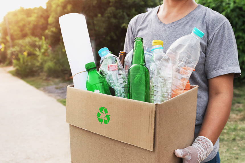 Persona con guantes sosteniendo una caja de cartón con botellas de vidrio y plástico para reciclaje, en un entorno al aire libre. 